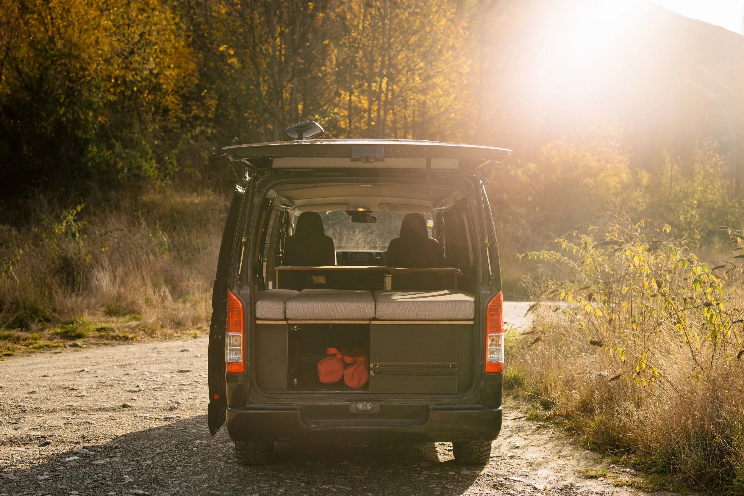 View of the open back of a black van parked on a dirt trail in a forest with fall foliage, with the sun shining brightly.