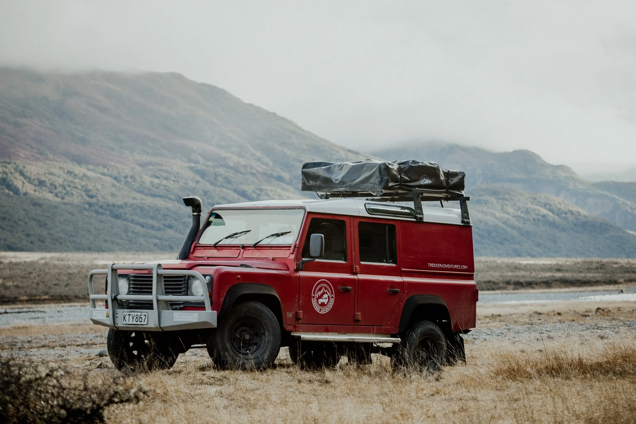 Red off-road vehicle with roof cargo carrier parked on grassy terrain with mountains in the background.