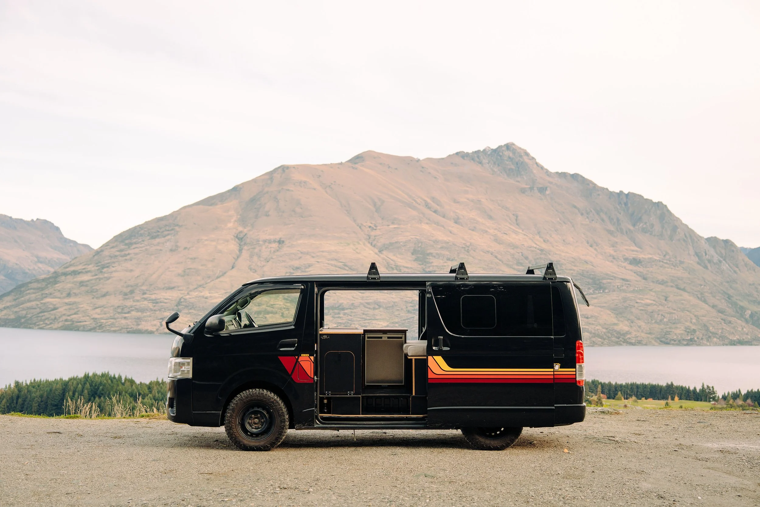 Black camper van parked in front of a mountain landscape, with a lake and trees in the background.