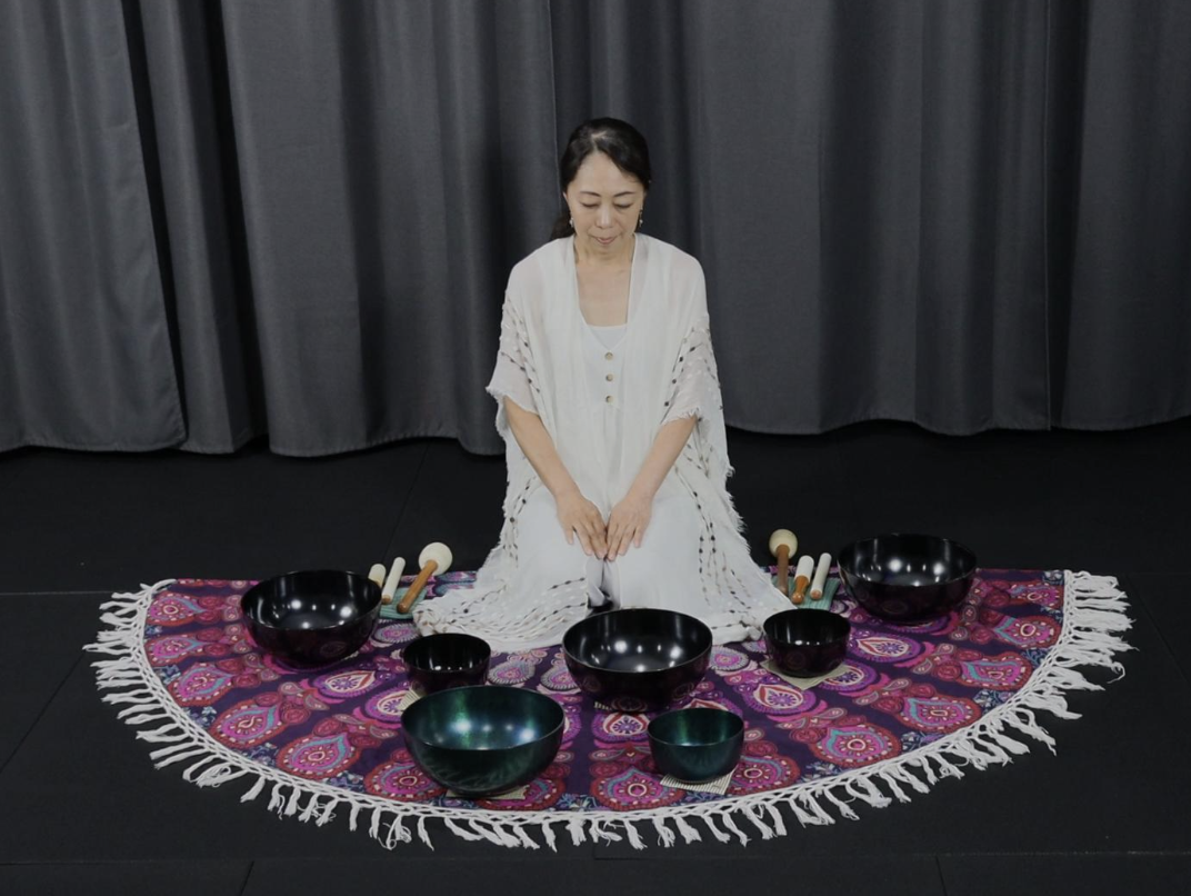 Woman sitting on a colorful fringed cloth with singing bowls around her on a stage with gray curtains behind.