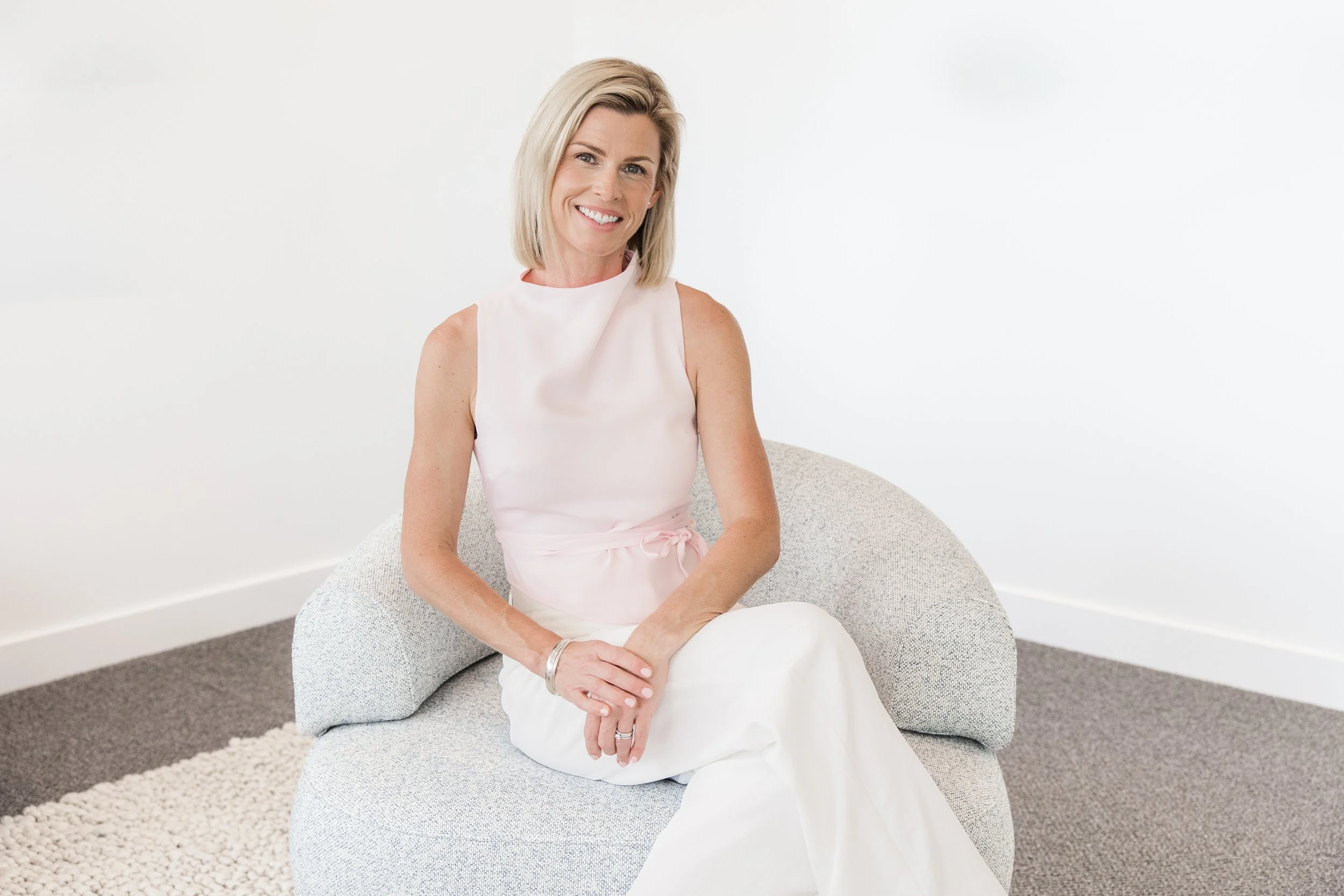 A smiling woman with blonde hair sitting on a light gray textured chair in a white room.