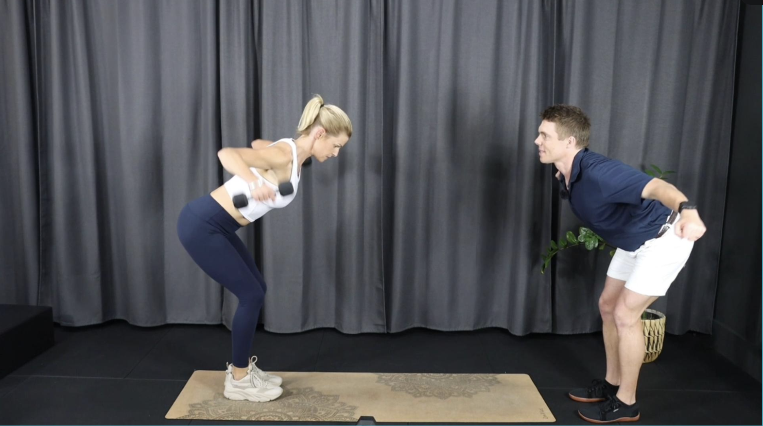 A woman performing bent-over rows with dumbbells supervised by a man in an indoor setting with dark curtains and a potted plant.