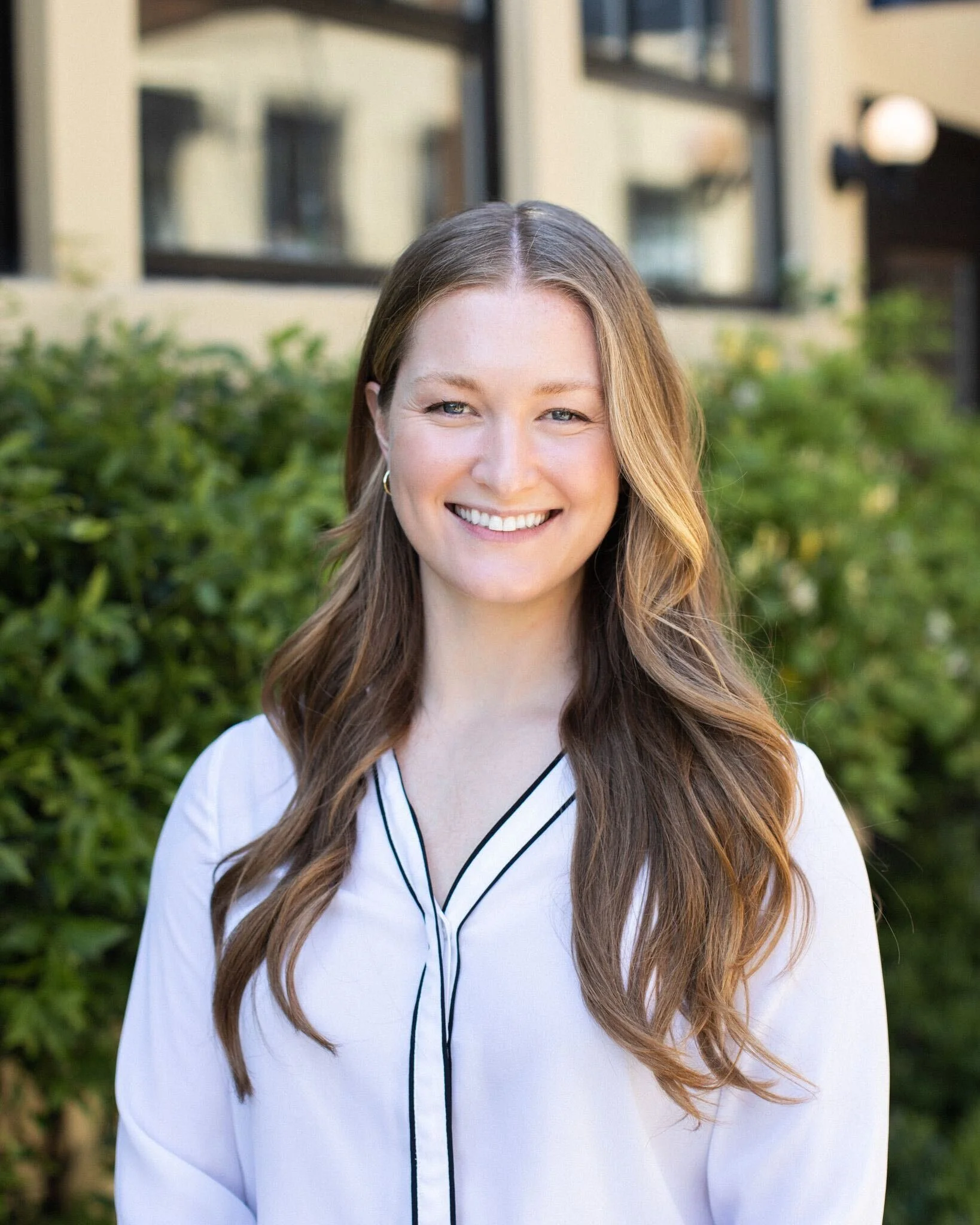 A woman with long, wavy brown hair, smiling outdoors in front of green bushes and a building with large windows.