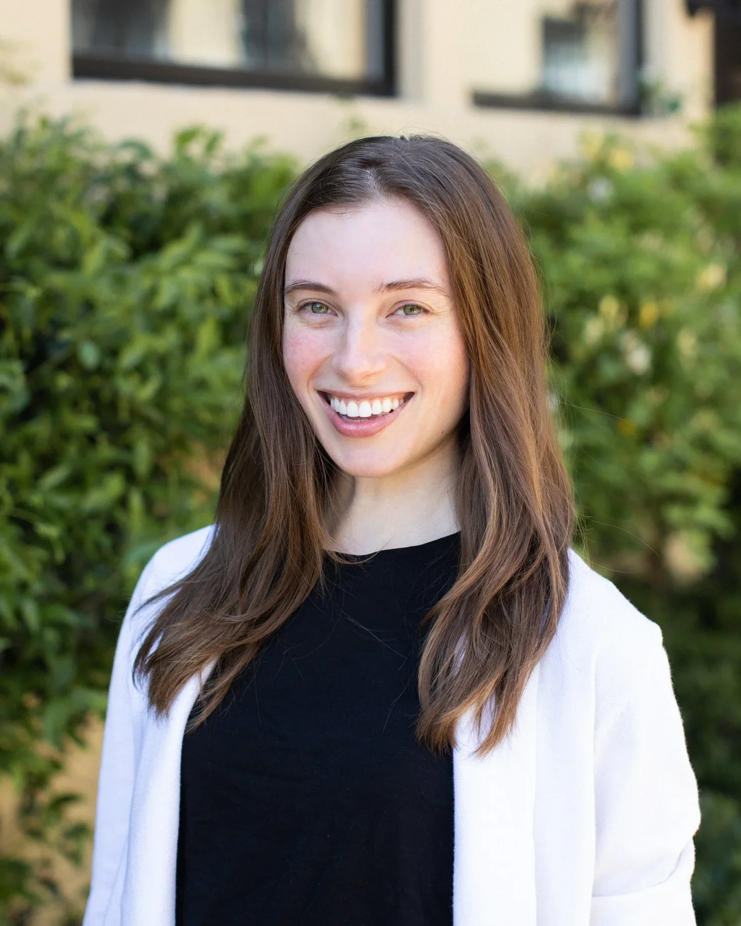 A young woman with long brown hair, wearing a black top and white jacket, smiling outdoors with green foliage and a building in the background.