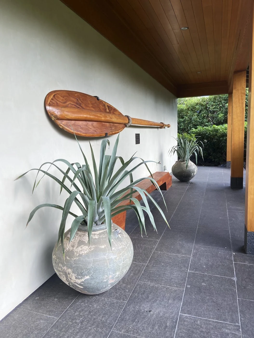Outdoor corridor with large potted plants, wooden bench, and a wooden paddle sculpture mounted on the wall.