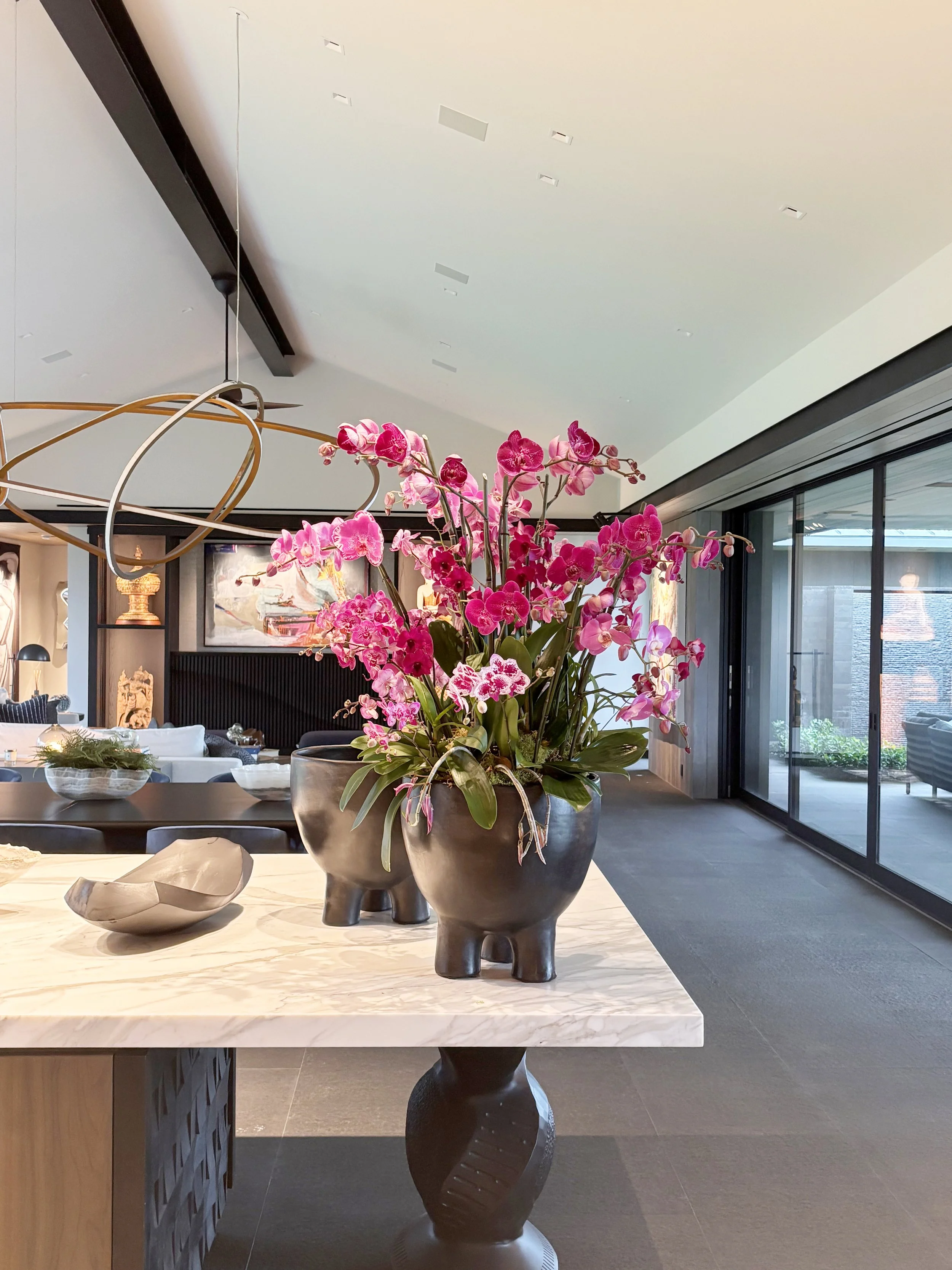 A large, vibrant pink orchid centerpiece on a black, sculptural vase sits on a marble table in a modern home along the Kona coast.