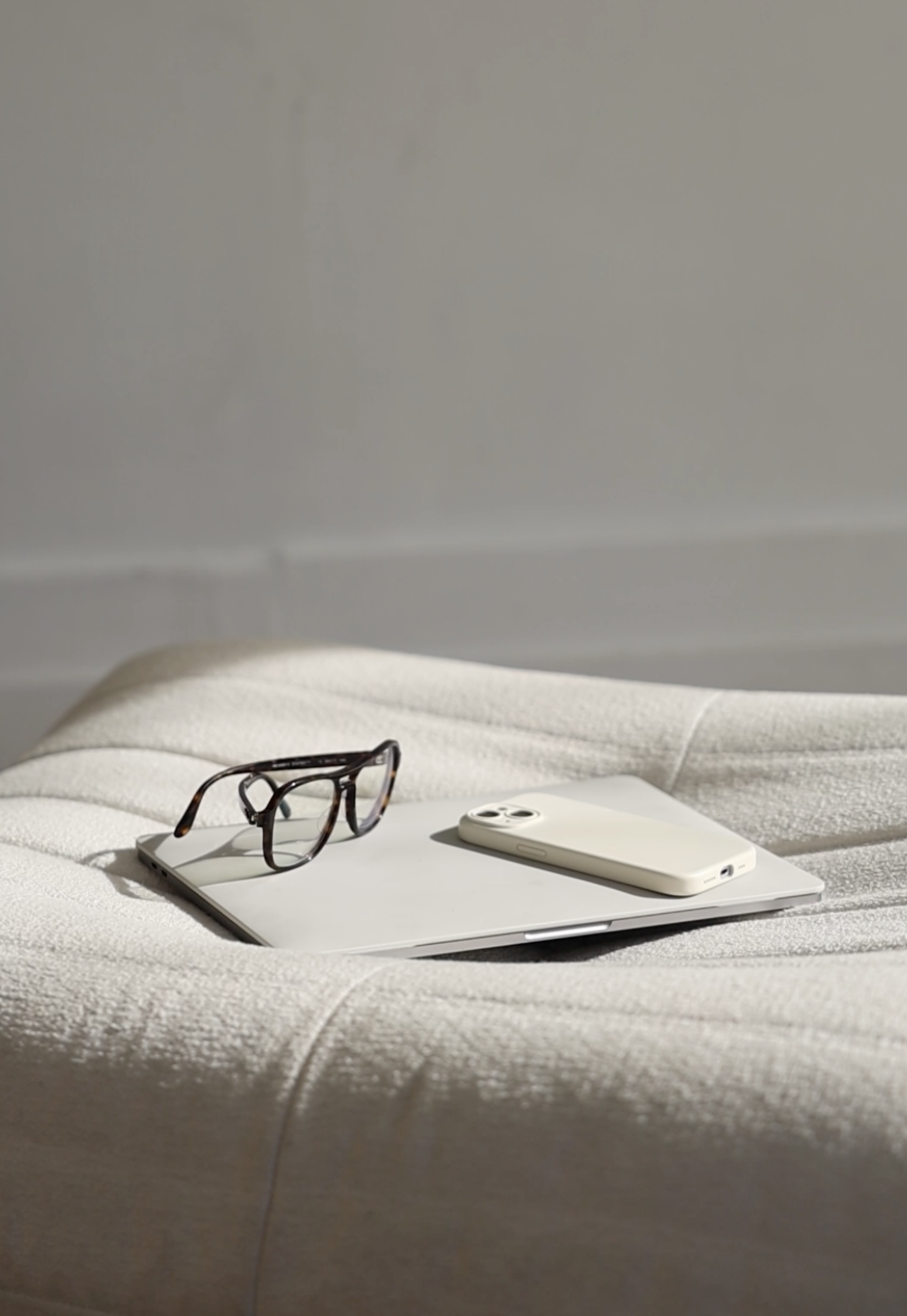 A laptop and phone displayed on a white lounge with a pair of reading glasses.