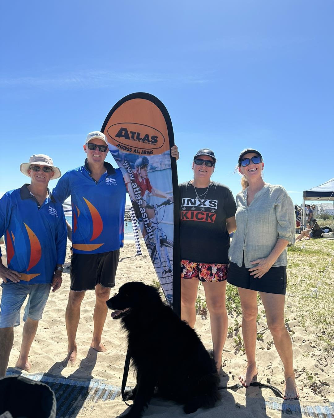 Four women and one man standing on a sandy beach next to a flotation device with the logo 'ATLAS' at a sunny outdoor event. They are smiling and wearing casual summer clothes, sunglasses, and hats. A large black dog is sitting in front of them.