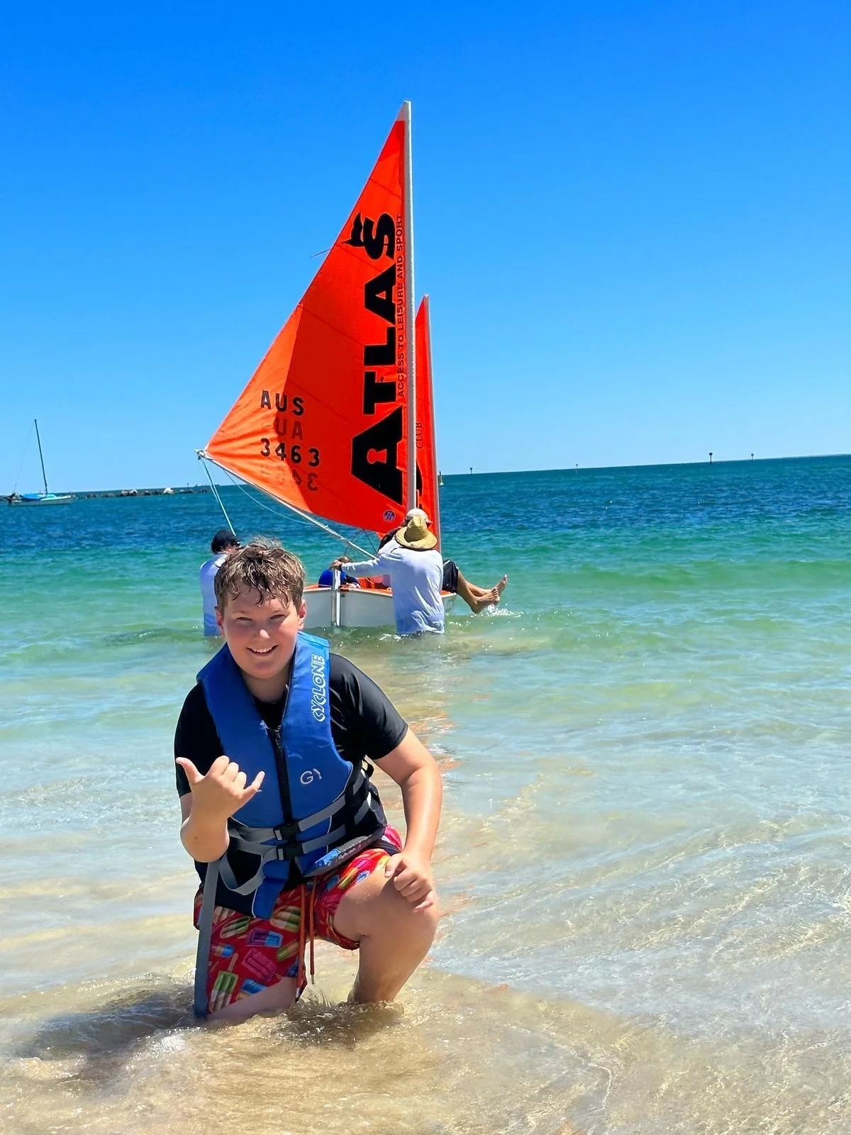 A cheerful boy kneeling in shallow water at the beach, making a shaka sign. In the background, people are preparing a small sailboat with a bright orange sail. The scene is sunny with clear blue skies and the ocean extending to the horizon.