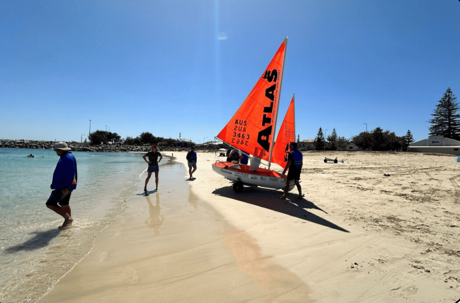 People stand on a sandy beach next to a small sailboat with bright orange sails. The people are dressed in casual beachwear, and some are walking in the shallow water. The scene is under a clear blue sky with a few trees in the background.