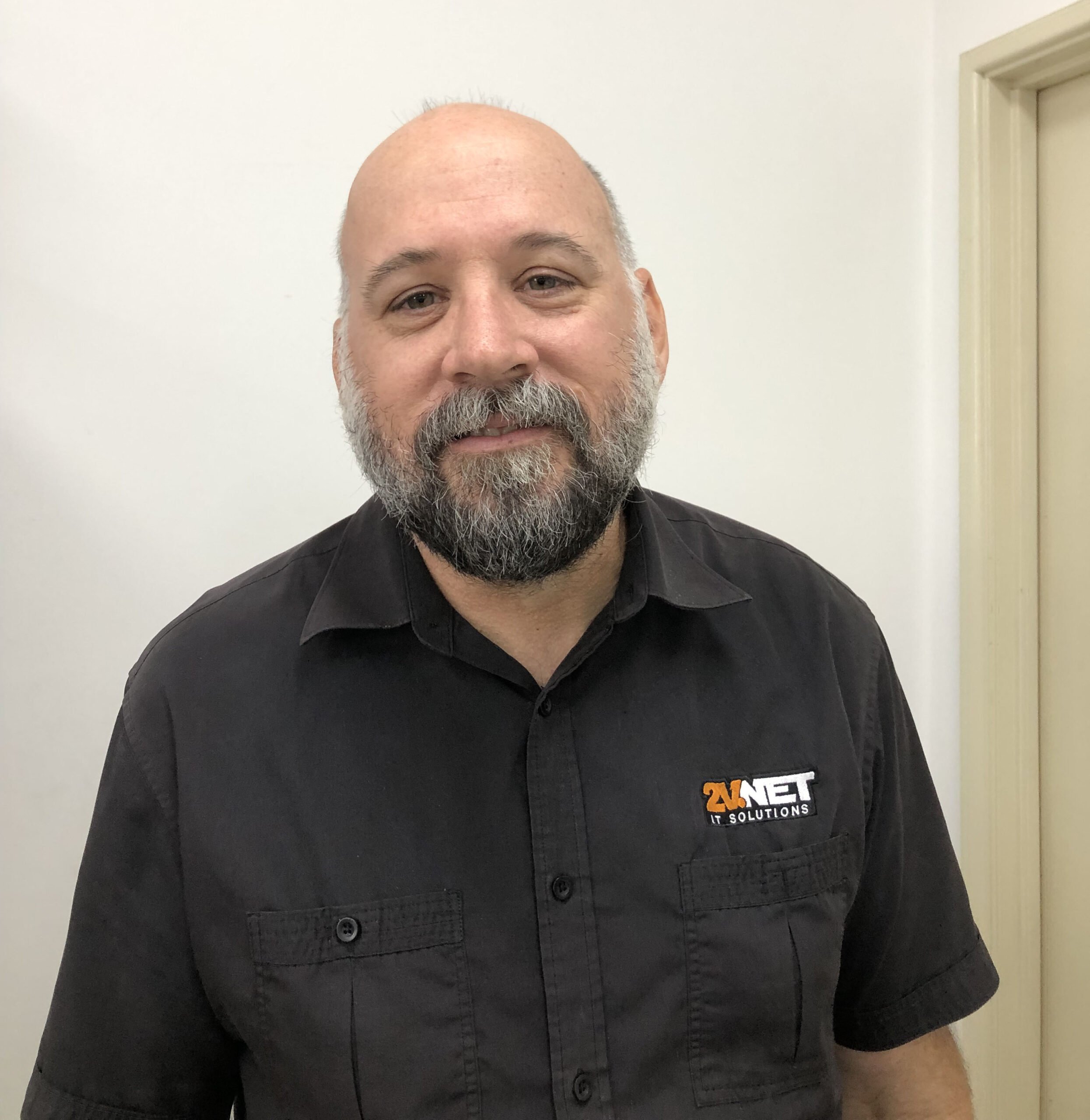 Andrew Toovey, wearing a black shirt with a logo that reads '2V NET IT SOLUTIONS,' standing against a plain wall.