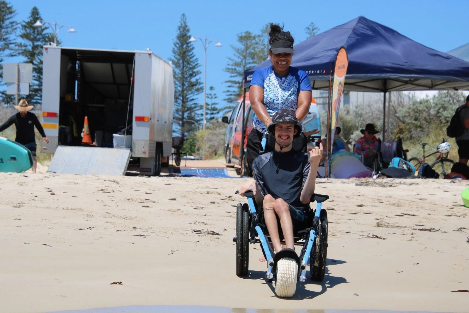 A woman pushing a man in a wheelchair on a sandy beach, smiling. There are tents and people in the background.