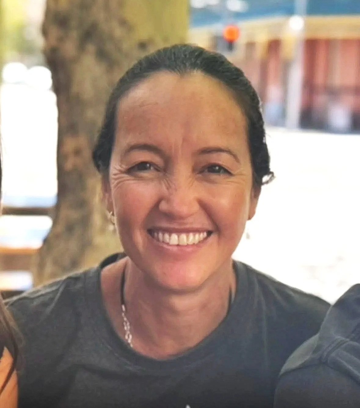 Kelly Bennett wearing a black shirt, outdoors with a blurred background of trees and buildings.