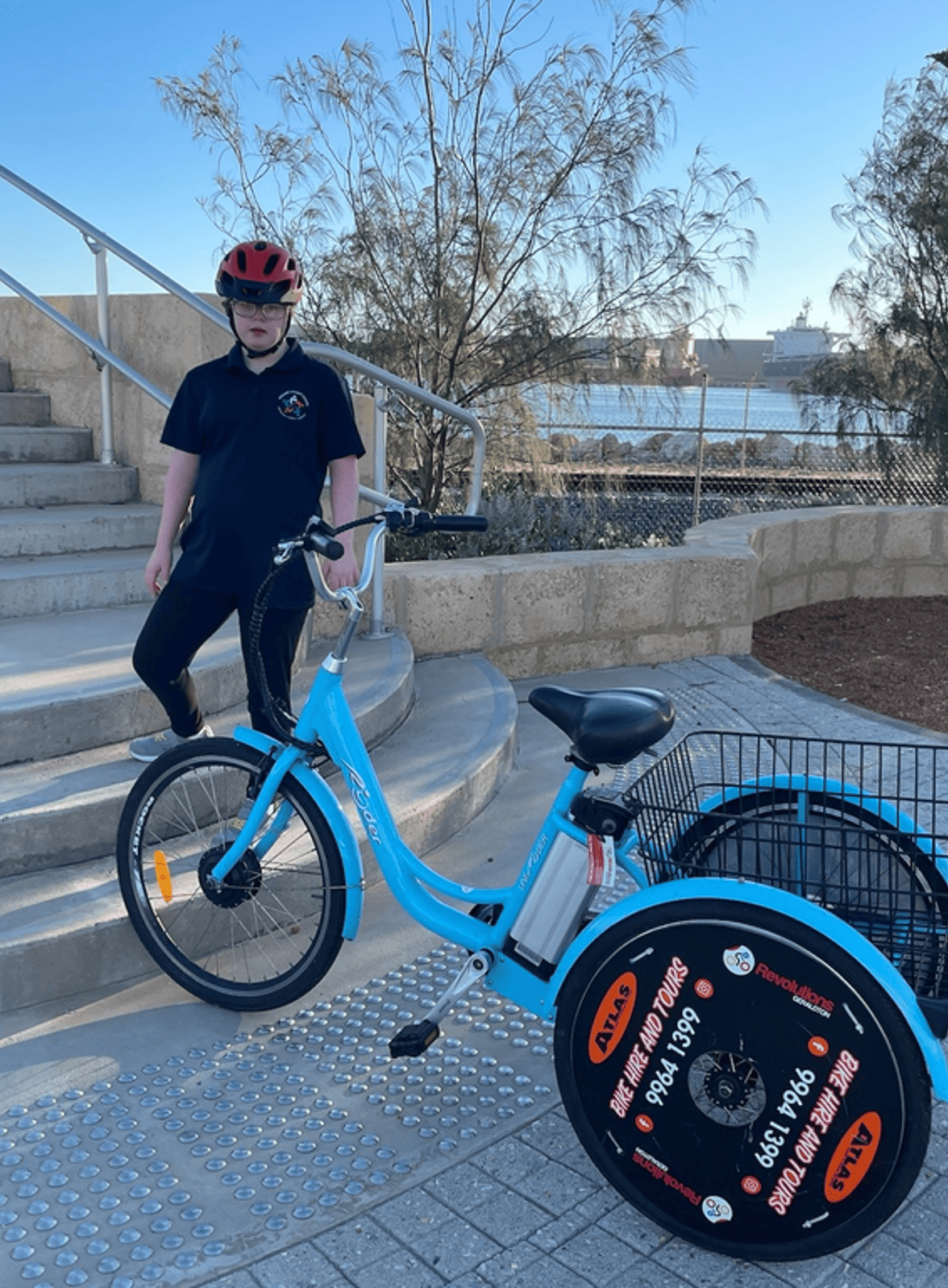 A young boy wearing glasses, a helmet, and a navy polo shirt, stands next to a blue bike with a front wheel and two large rear wheels for bike tours, near concrete stairs, with trees, water, and ships in the background.