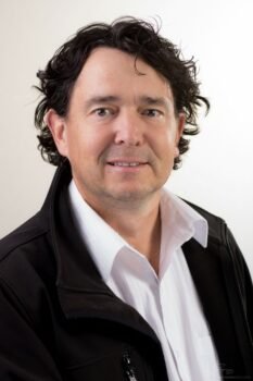 Adam Murszewski, wearing a black jacket over a white shirt, smiling at the camera.