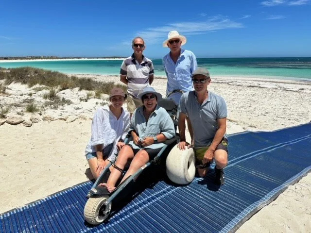 A group of five people, including children and adults, with the ocean in the background. One person is using a beach wheelchair and accessing the beach via a pathway over sand for wheelchairs and strollers.