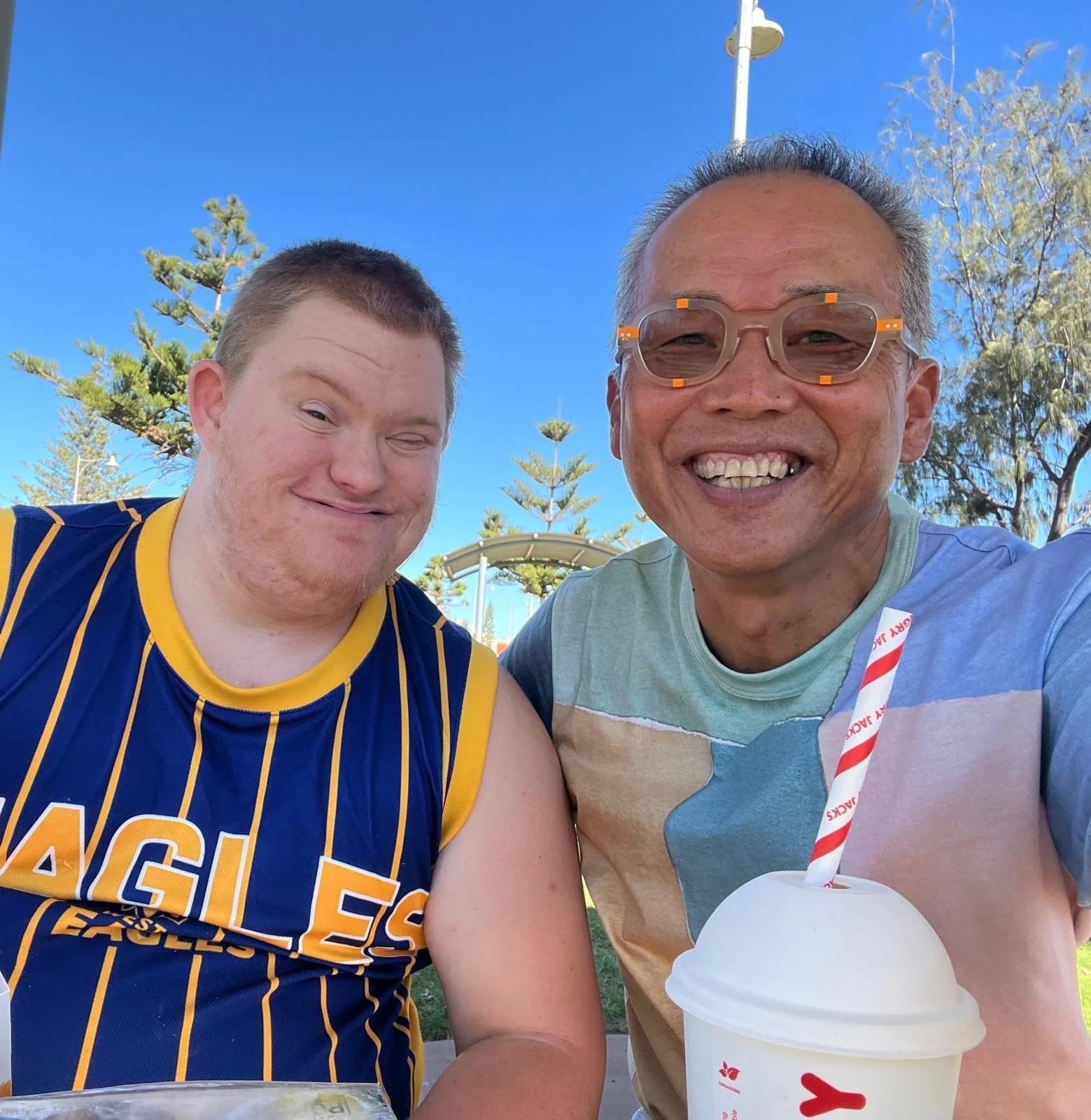 Two smiling men taking a selfie outdoors, one wearing glasses and a casual t-shirt, the other in a sleeveless sports jersey, with trees and a blue sky in the background.