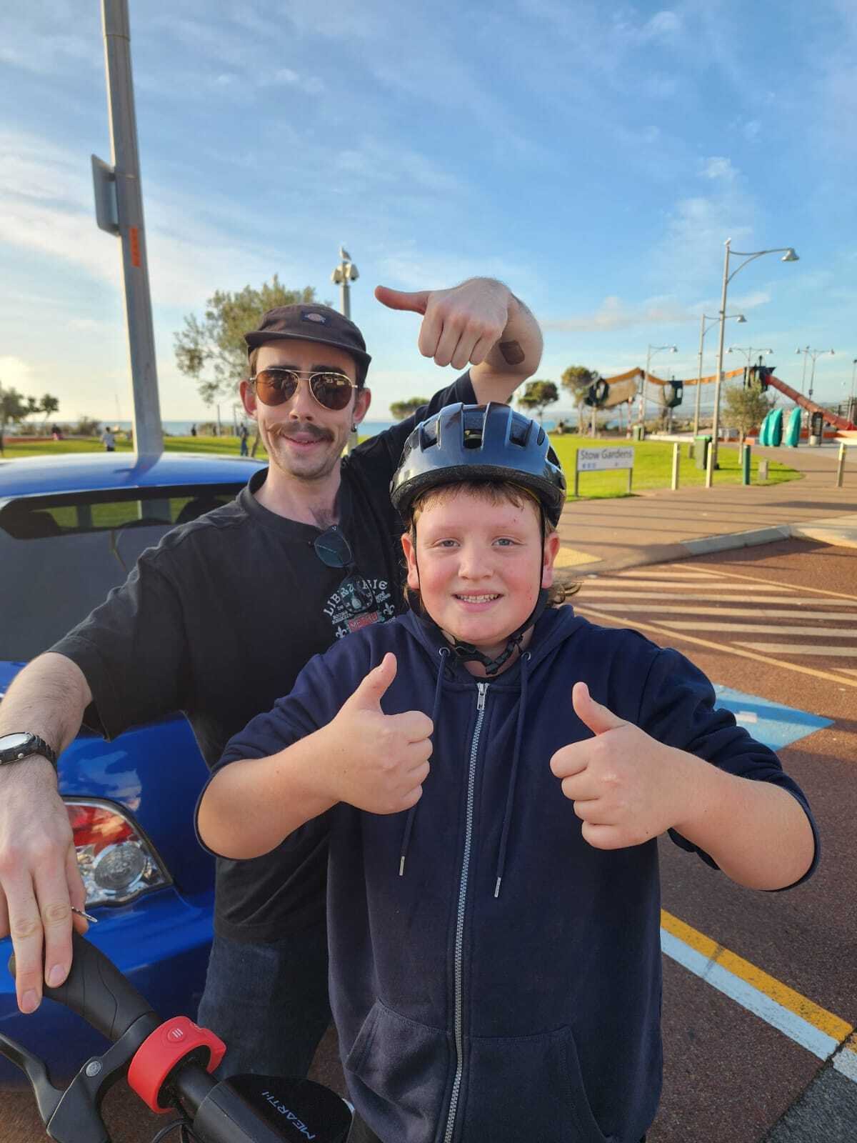 A man and a boy wearing helmets are smiling and giving thumbs up in a parking lot near a playground. The man is making a gesture with his hand above the boy's head, resembling a fist bump.