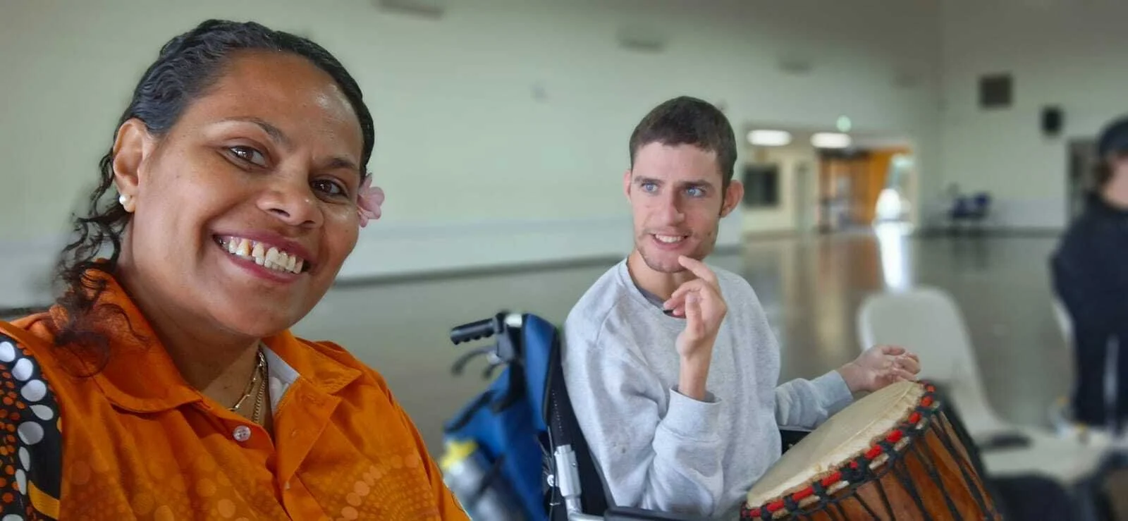 Smiling woman in orange shirt taking a selfie with a man in a wheelchair playing a drum in a room with blurred background.