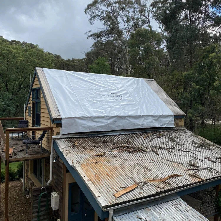 Two sections of a house roof with different roofing materials, one under construction with a plastic covering, and the other covered in rusty corrugated metal with debris, surrounded by trees.