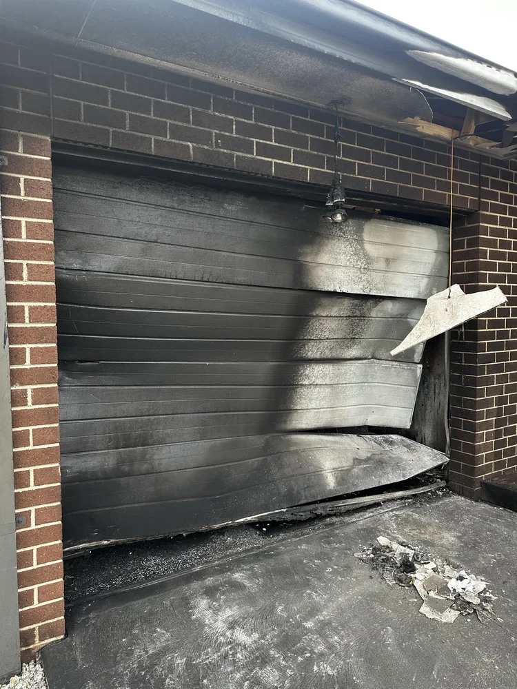 A burned garage door with smoke damage and a fallen, damaged security light, showing signs of fire damage.