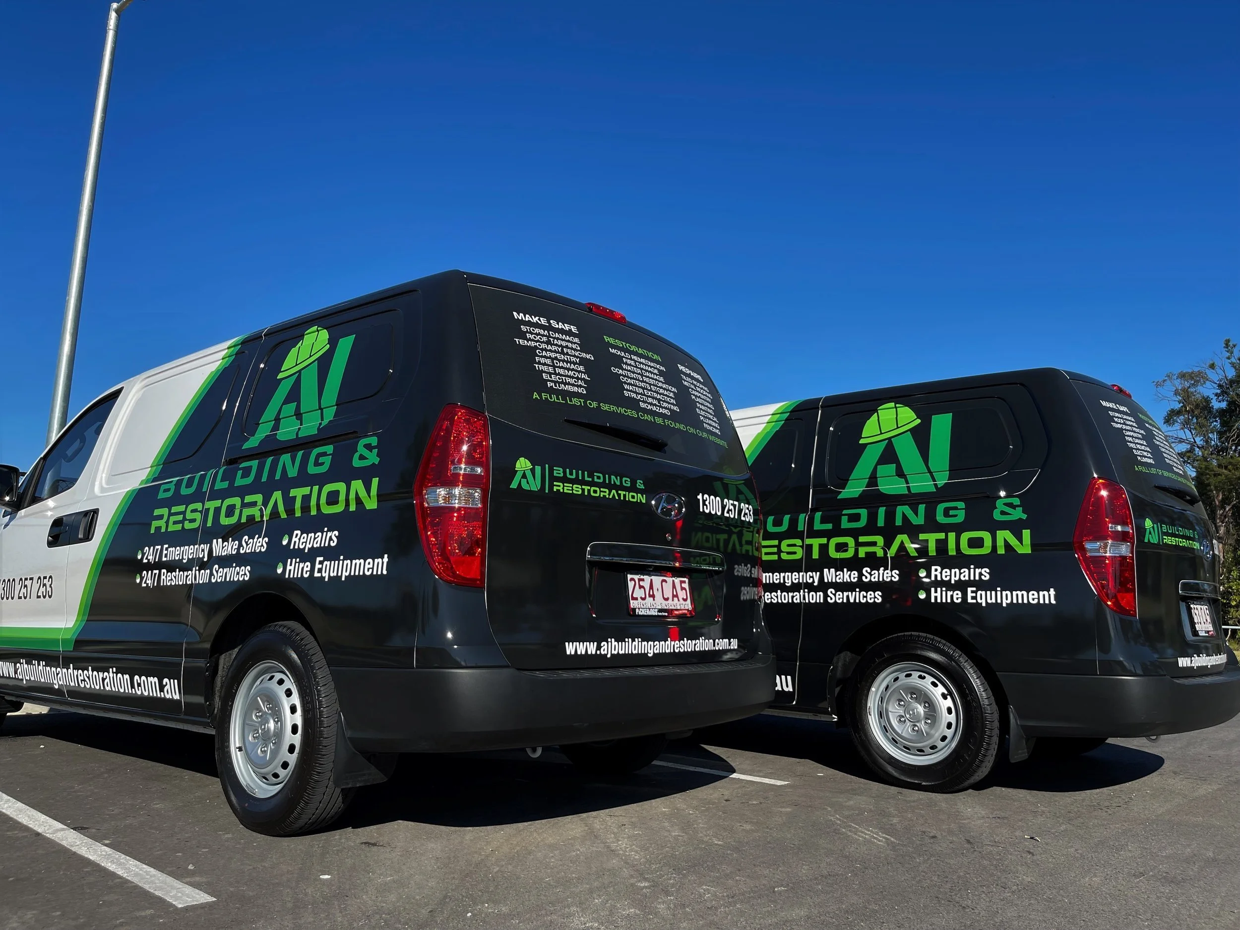 Two black and white service vans with green and white branding for a building and restoration company, parked outdoors under a clear blue sky.