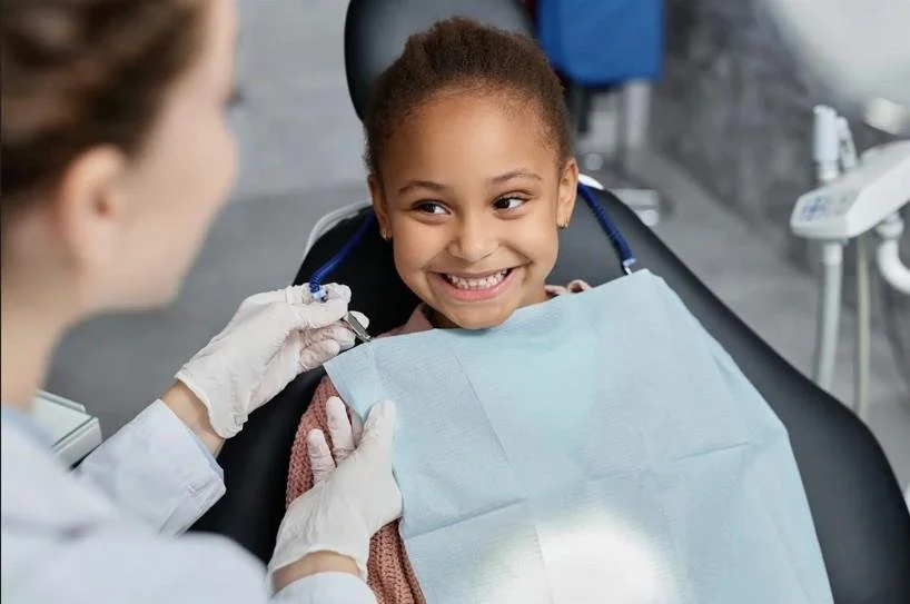 Smiling young girl sitting in a dental chair as a pediatric dentist prepares for a tooth-colored filling — gentle, natural-looking cavity treatment for kids at Starlight Dental.