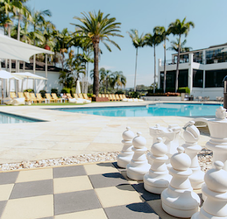 Poolside scene with large outdoor pool, palm trees, pool chairs, and oversized chess pieces on a checkered chessboard.