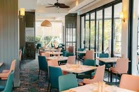 Interior of a modern restaurant with tables and chairs, large windows letting in natural light, and a view of trees outside.