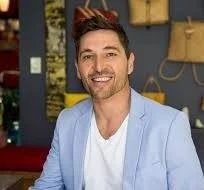 A man with dark hair and a beard, wearing a light blue blazer and white shirt, smiling indoors with a colorful background.