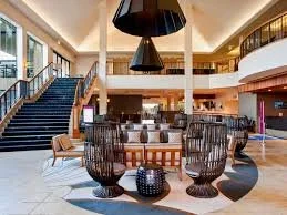 Luxurious hotel lobby with a round wooden dining table, four black decorative chairs, and adjacent staircase, with large windows and modern lighting fixtures.