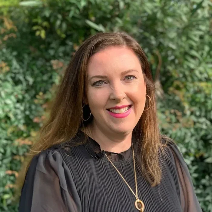 A woman with long brown hair, wearing a black top with pleats, gold hoop earrings, and a gold necklace, smiling outdoors in front of green foliage.