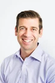 A smiling man with short brown hair wearing a light blue collared shirt against a plain white background.