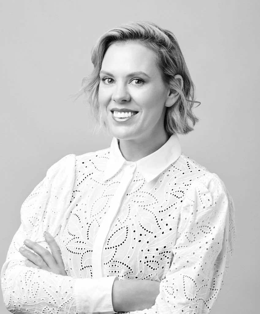 Black and white portrait of a woman with short wavy hair, smiling, wearing a white embroidered blouse, arms crossed.