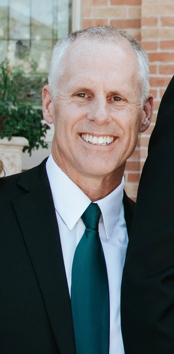 Close-up of a smiling middle-aged man wearing a black suit, white dress shirt, and green tie, standing outdoors with a brick building and plants in the background.