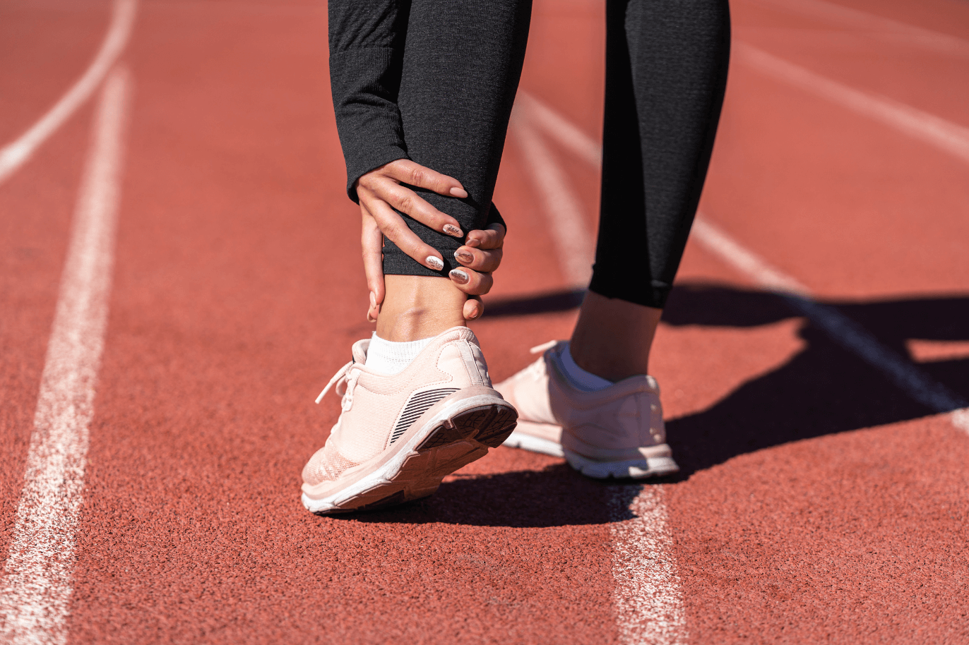 A person on a running track stretching, wearing pink running shoes and black athletic leggings.