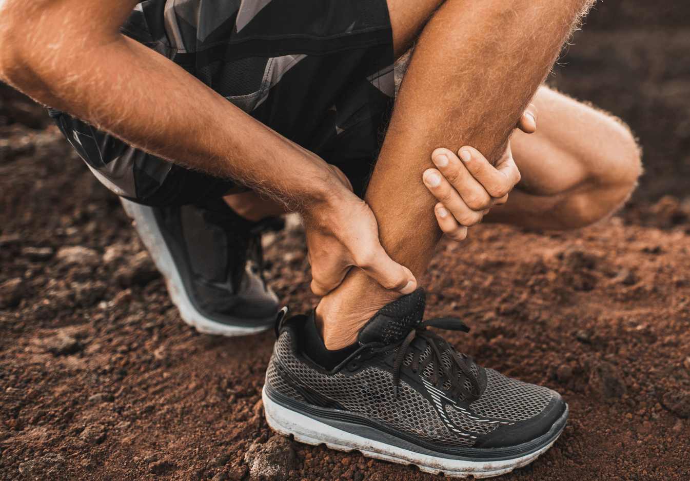 Close-up of a person in athletic shoes holding their ankle on rough ground, possibly experiencing injury or pain.