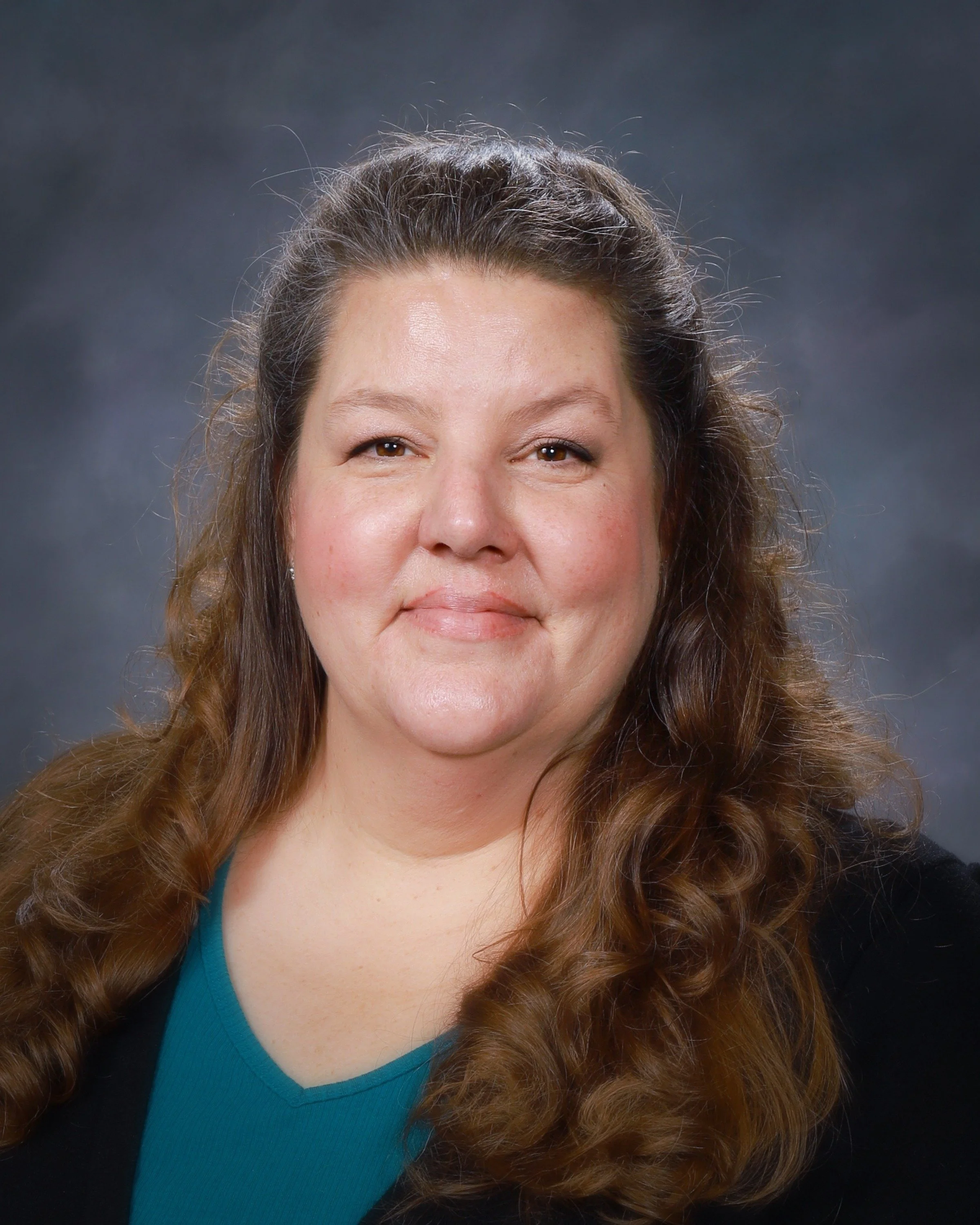 A woman with long brown hair smiling in front of a gray backdrop, wearing a gray T-shirt with yellow text and a graphic.