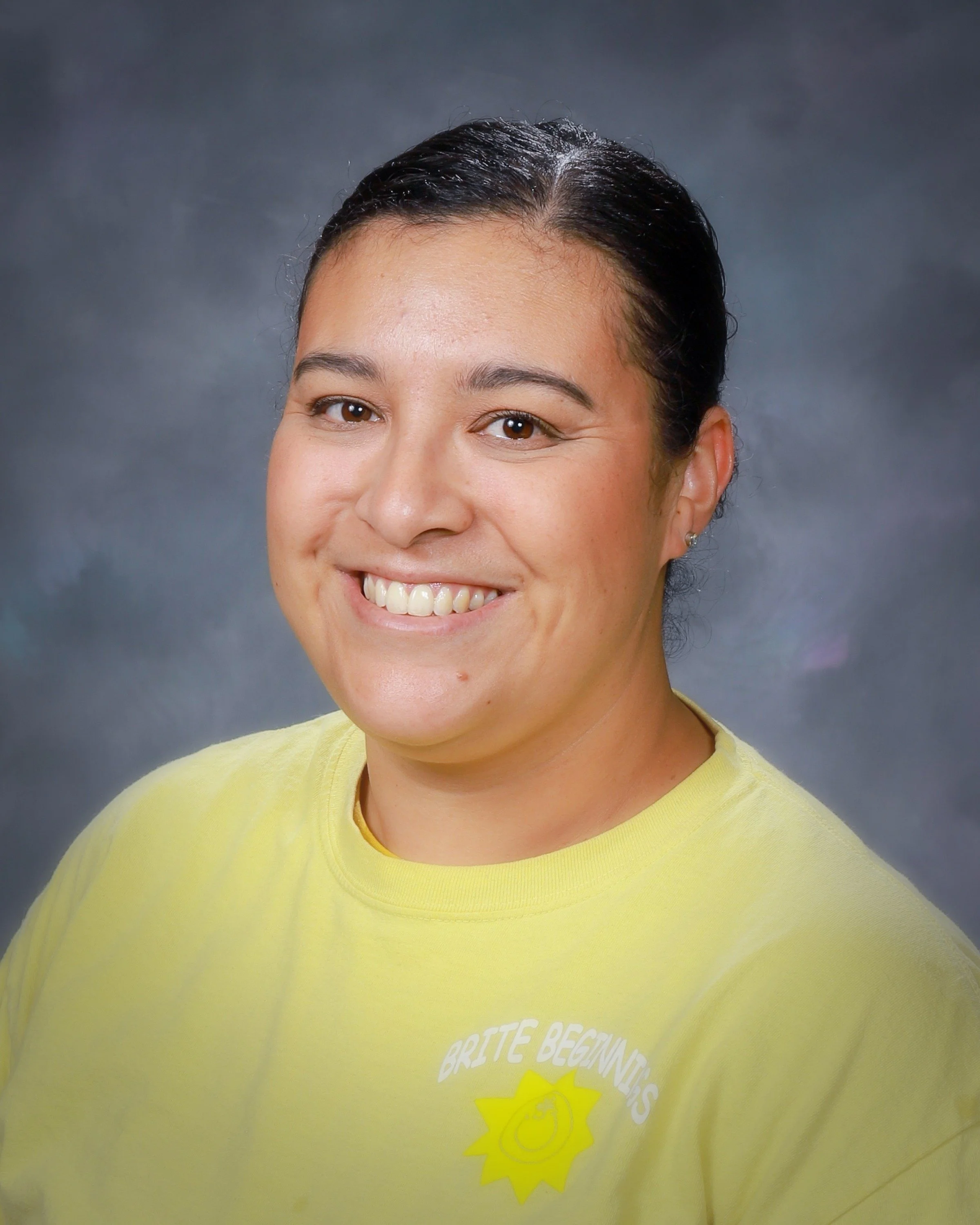 A woman with glasses and dark hair tied back, smiling, wearing a navy blue shirt with text and a sun logo, against a gray background.
