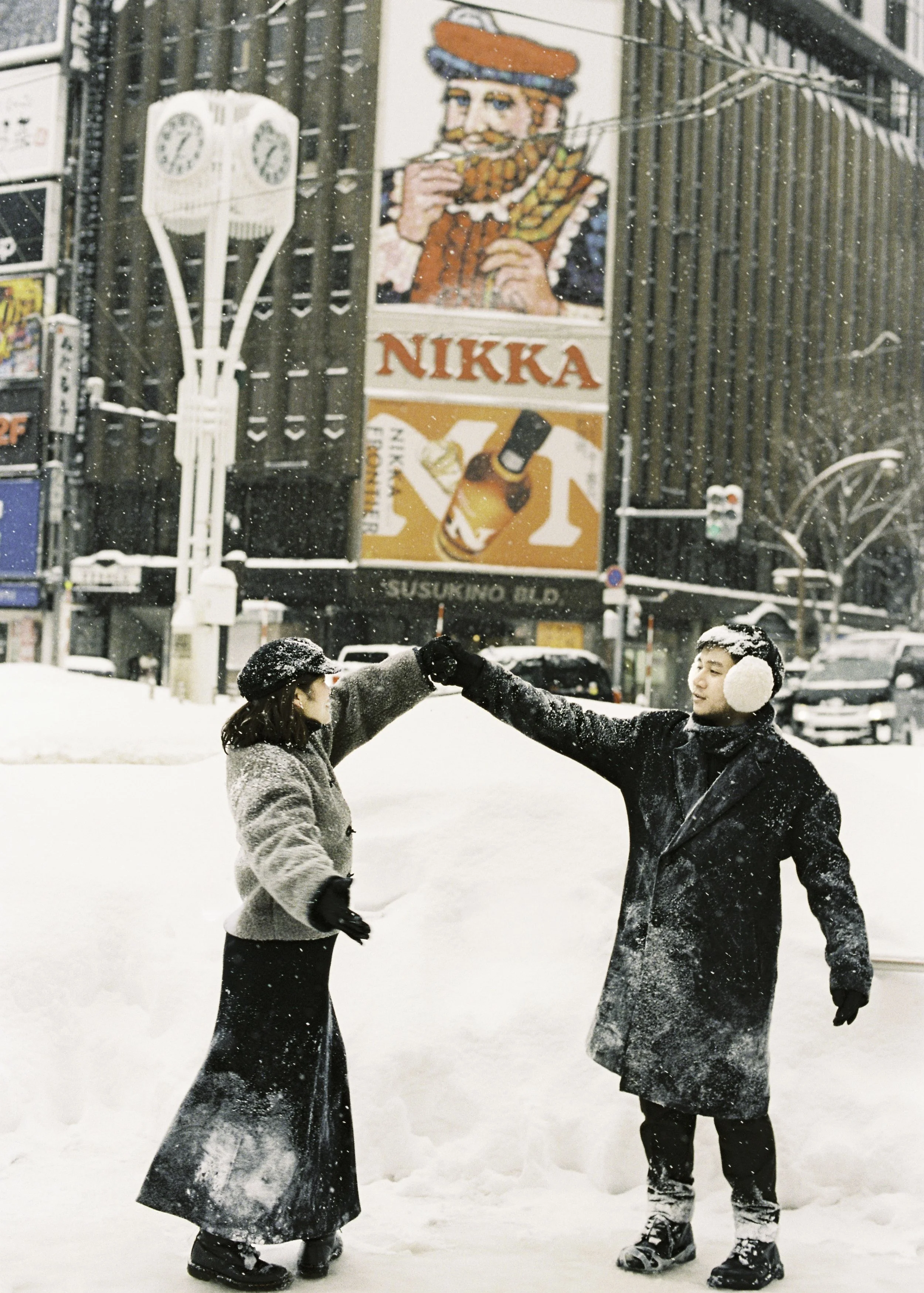 A man and woman in winter clothing exchange a fist bump in snow-covered city street with large building signs in the background. Couple photoshoot in Sapporo.
