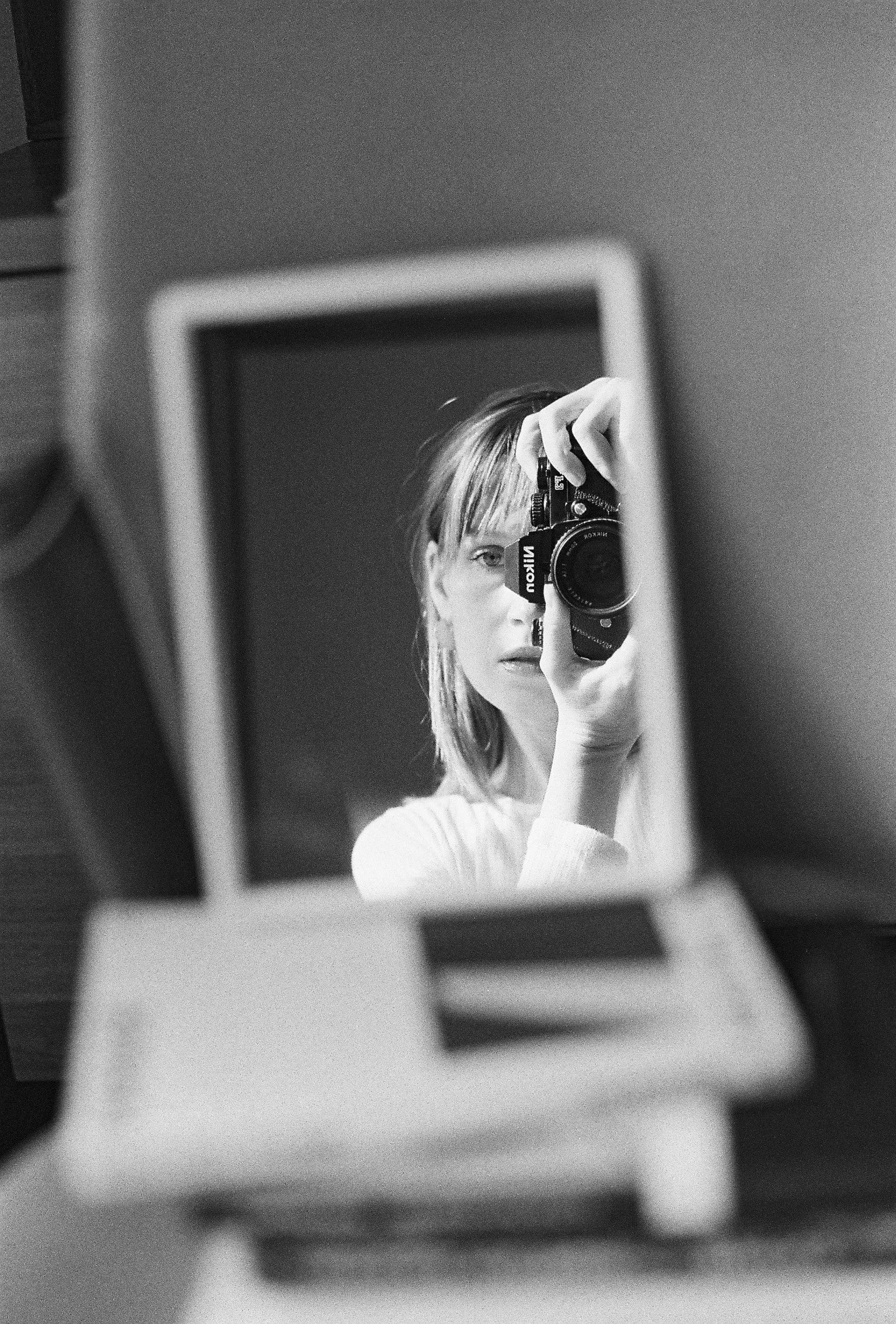 A woman taking a selfie using a Nikon camera, shown through her reflection in a mirror, in black and white.