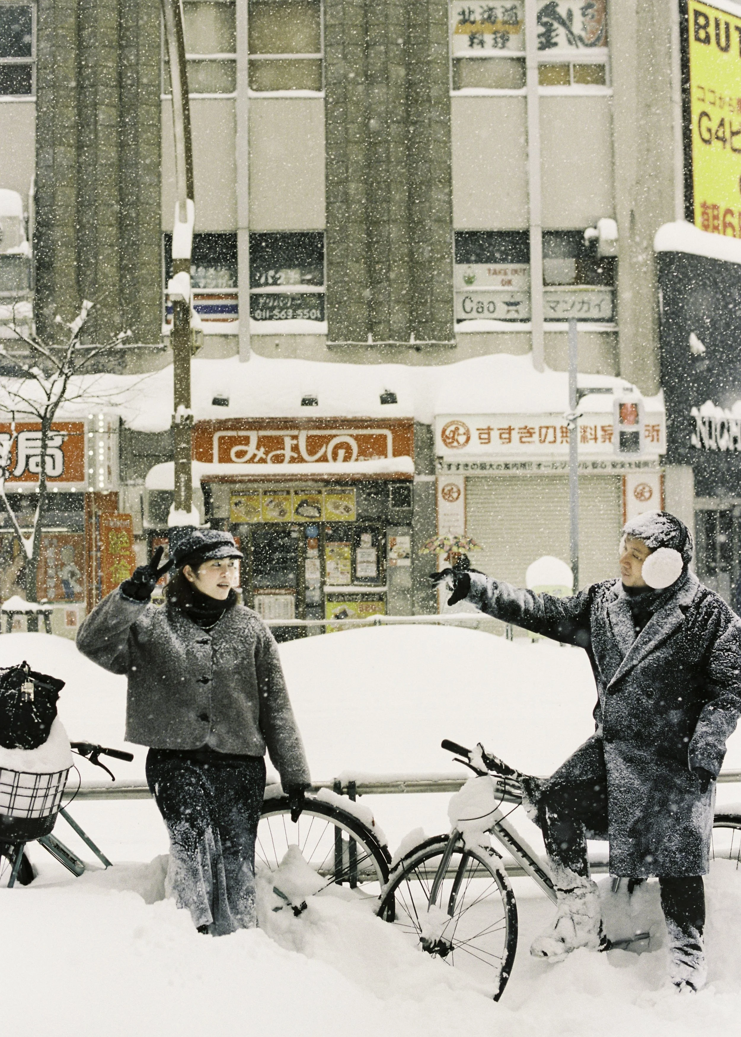 Two people dressed in winter clothing and earmuffs, standing with bicycles in a snow-covered city street during snowfall, engaging in conversation or greeting each other. Couple photoshoot in Sapporo.