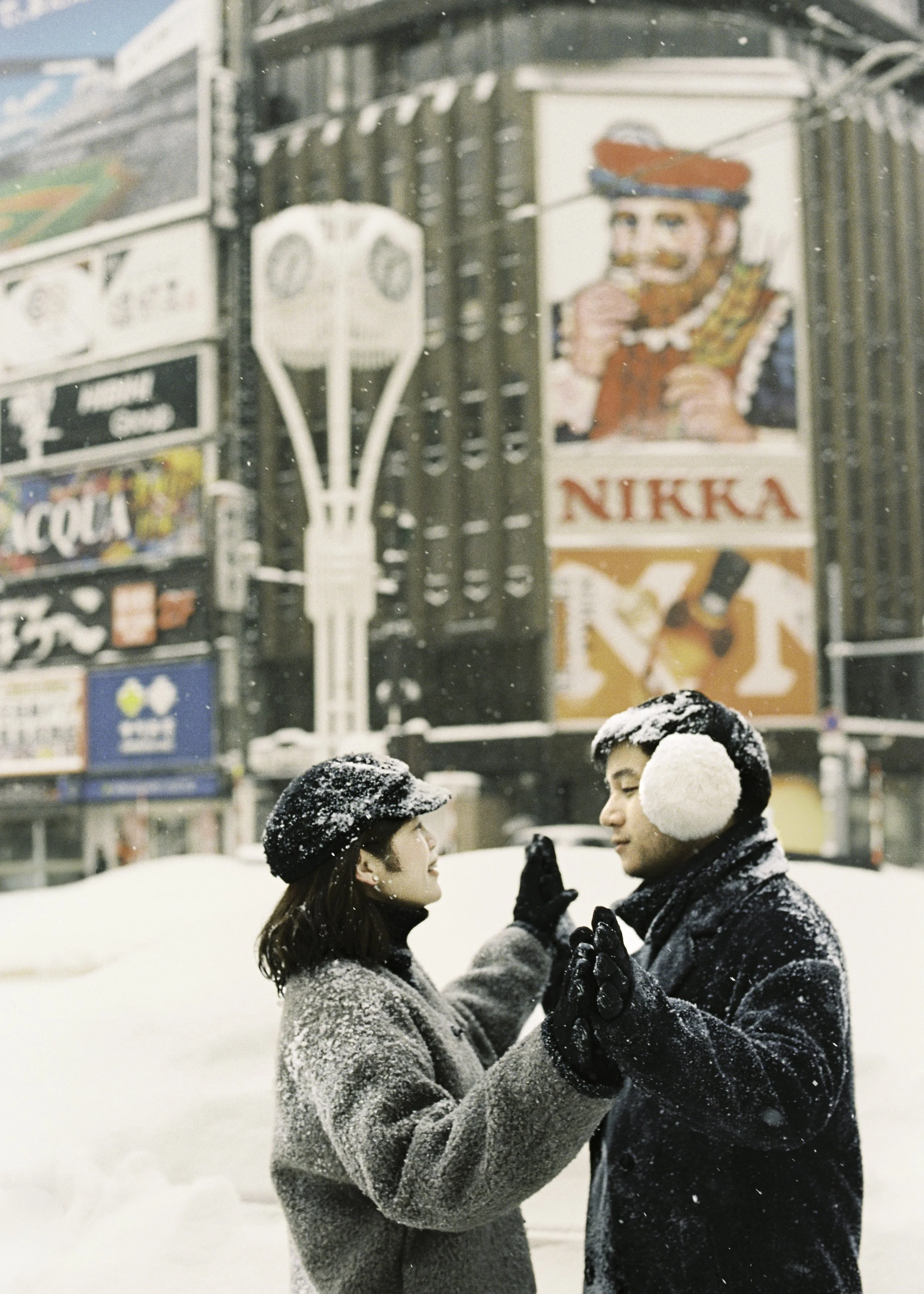 A couple wearing winter clothing and earmuffs standing close together in a snowy city, with colorful billboards and advertisements in the background. Couple photoshoot in Sapporo.