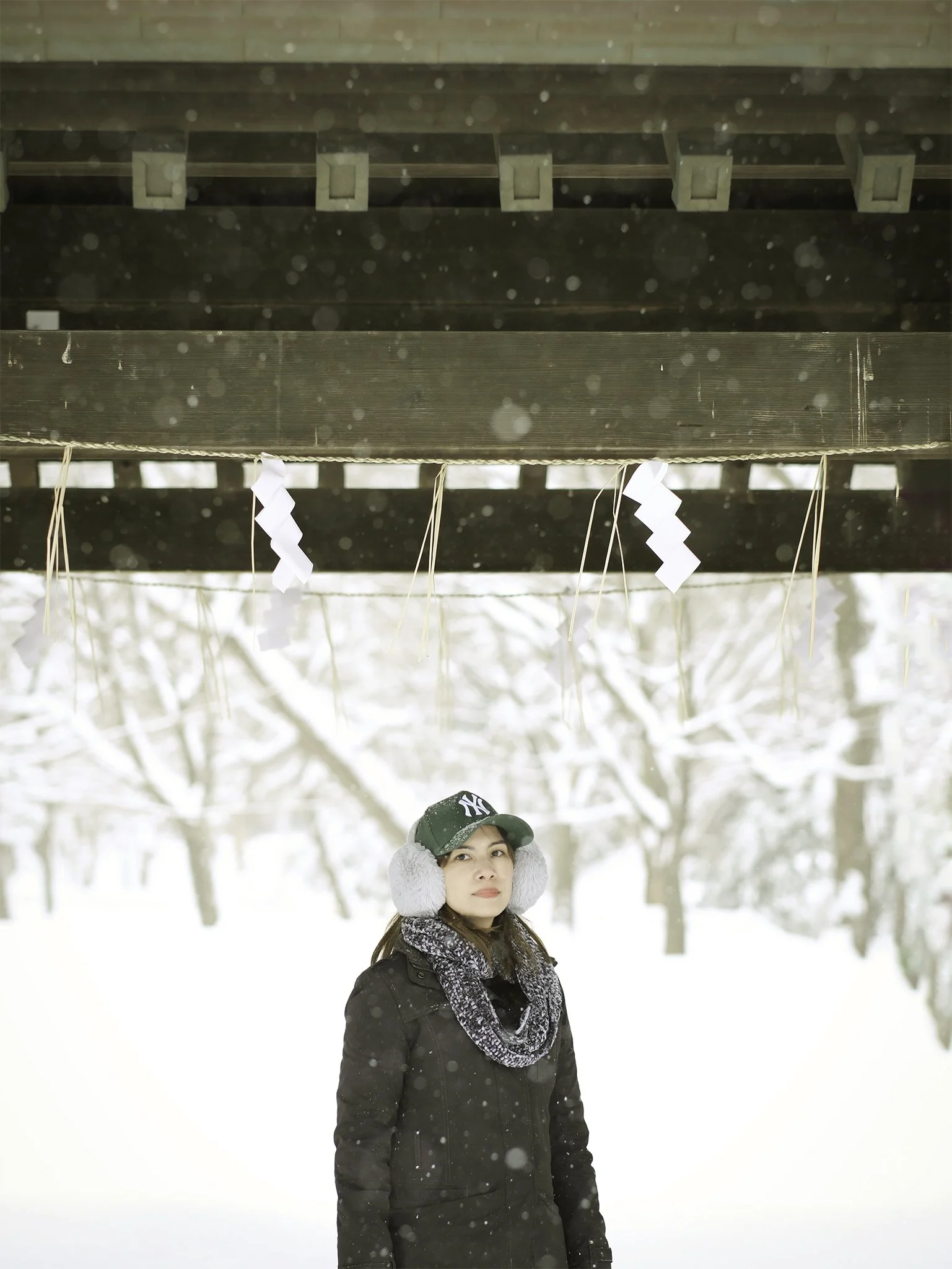 A woman standing outdoors in a snowy landscape, wearing earmuffs, a scarf, a black jacket, a cap, and snow falling around her, with a wooden structure above her decorated with paper and straw ornaments.
