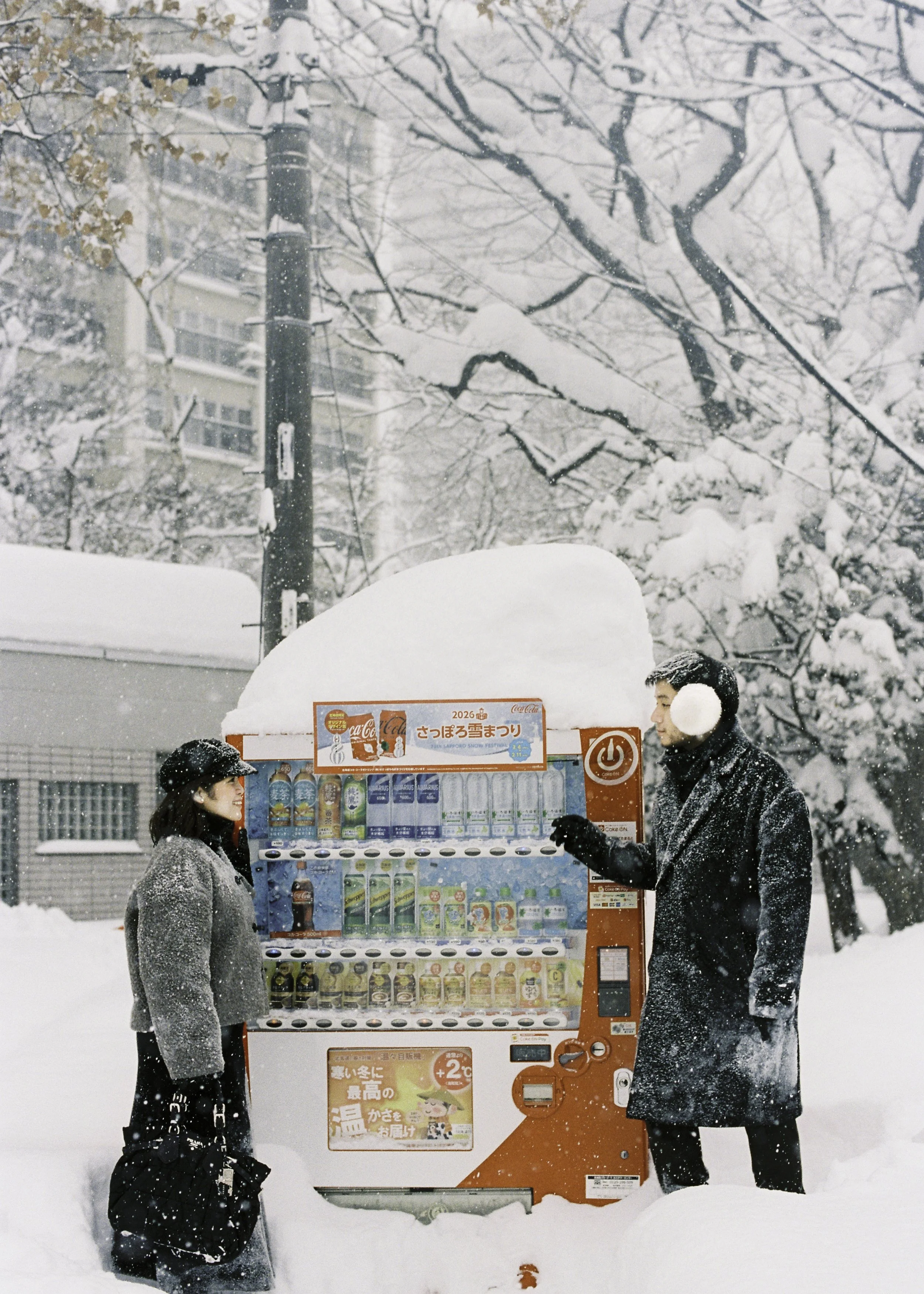 Two people in winter clothing and earmuffs stand at a vending machine in a snowy outdoor setting, with snow-covered trees and buildings in the background.