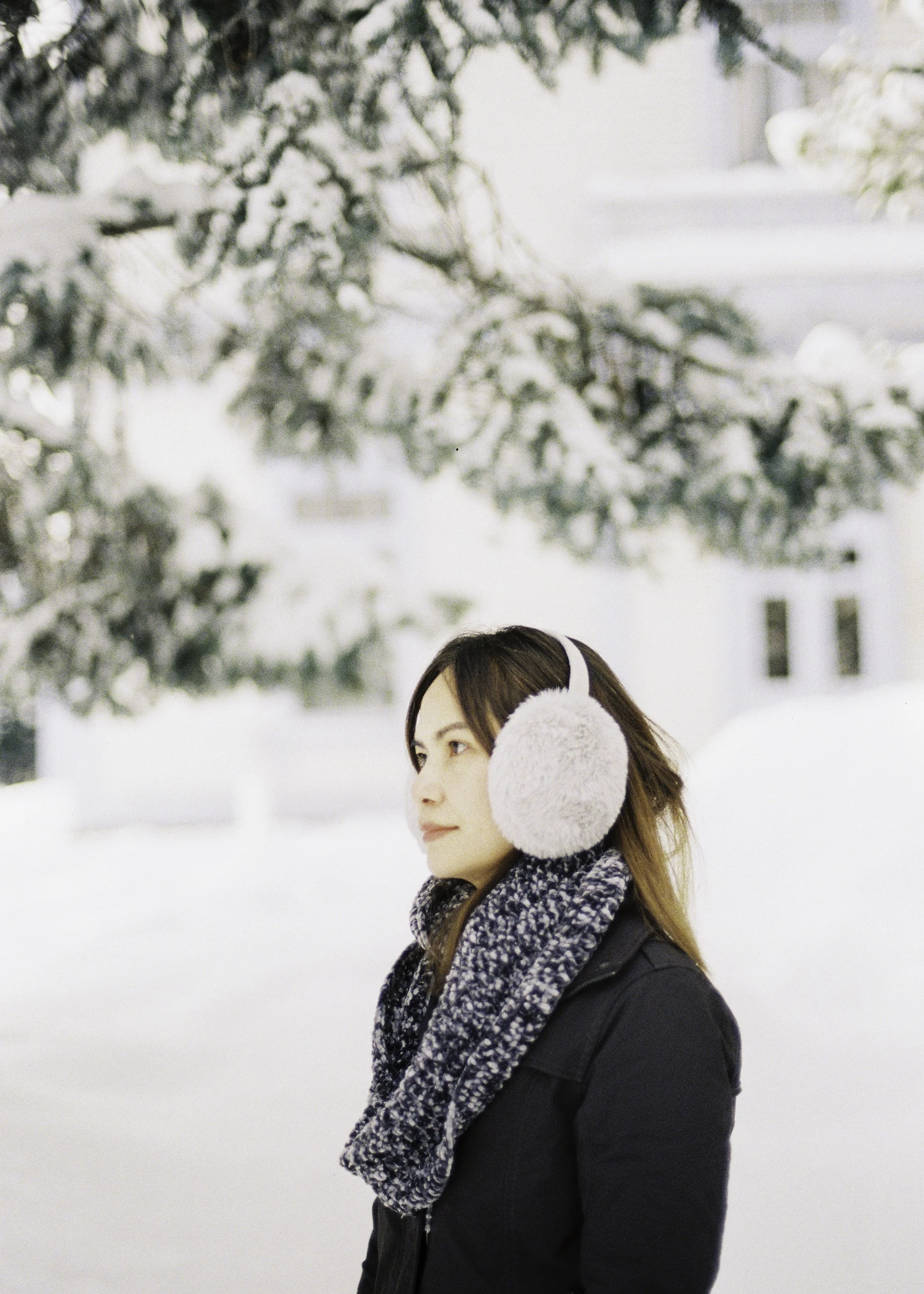 A woman wearing earmuffs, a scarf, and a winter coat standing outside in a snowy landscape with snow-covered trees background.
