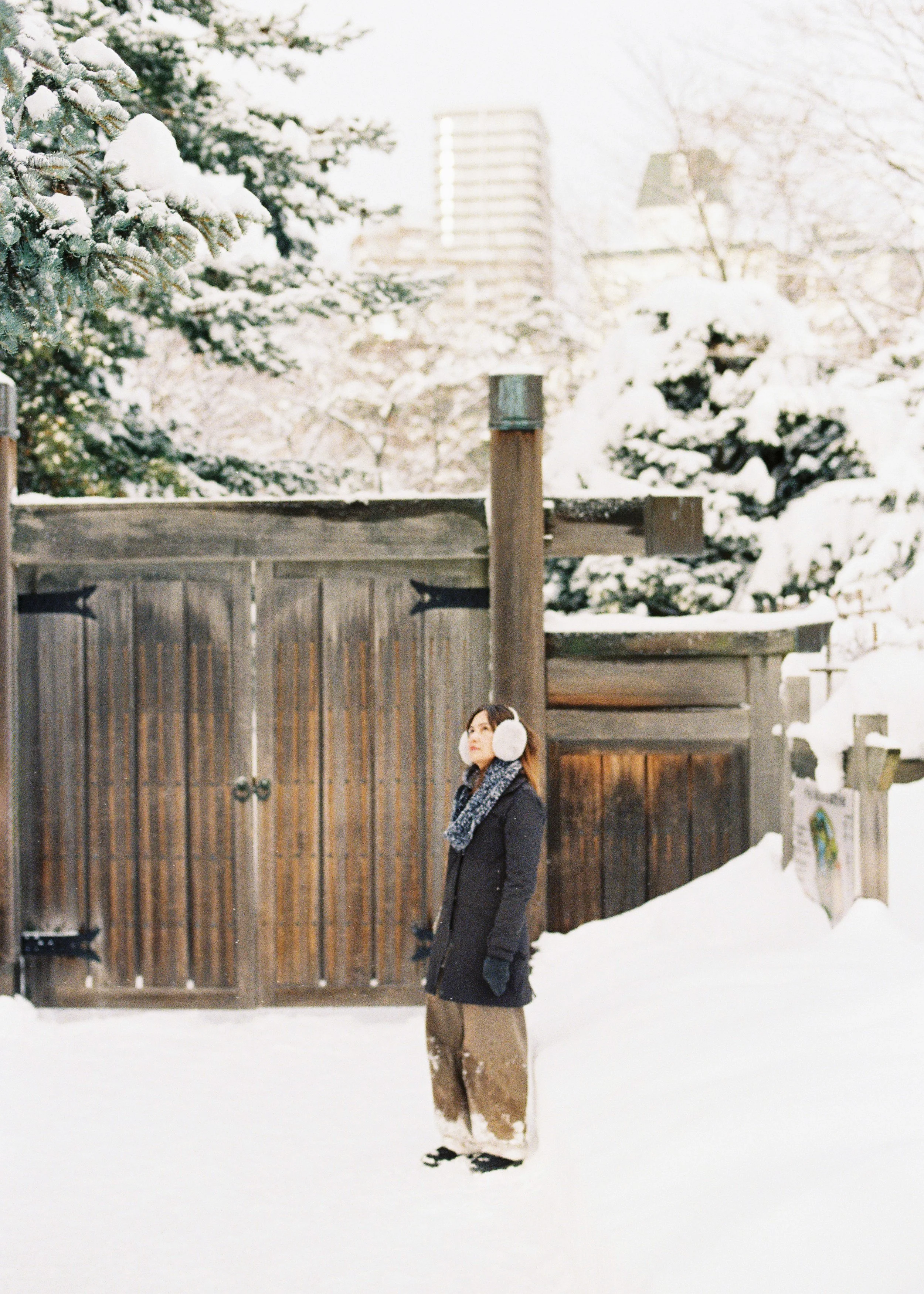 A woman standing in snow with headphones, dressed in winter coat, scarf, gloves, and snow boots, near a wooden fence and snow-covered trees.
