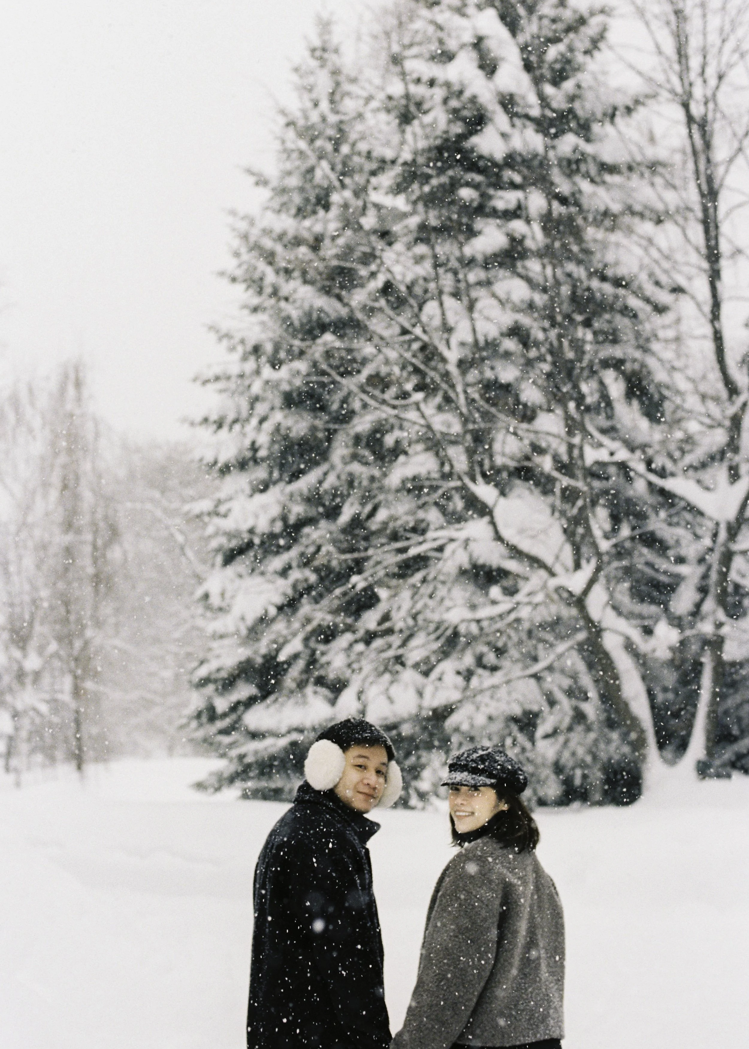 Two people standing in a snowy landscape with snow-covered trees in the background, smiling and dressed warmly for winter.