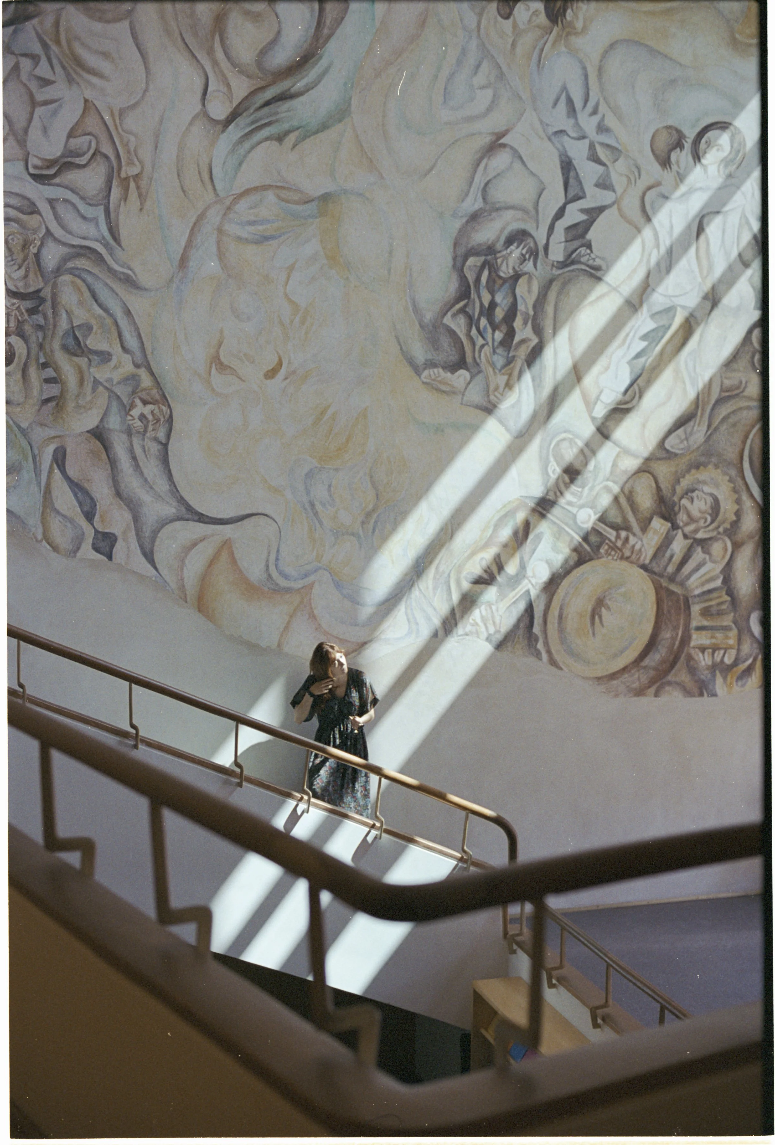 A woman in a floral dress stands on a staircase in front of a large abstract mural with swirling shapes and figures, illuminated by sunlight streaming through a window.
