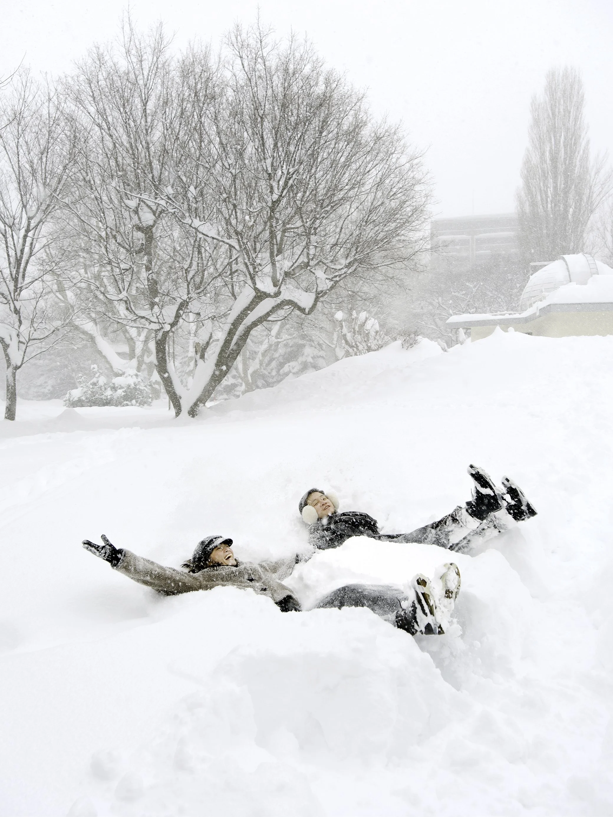 Two people lying in the snow, laughing and playing in a snowy landscape with leafless trees and a building in the background.